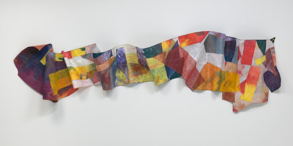 Sam Gilliam | Sam Gilliam: Sewing Fields | Friday 13 June  – Sunday 16 November 2025 | IMMA | Image: Sam Gilliam |  photo of a horizontal strip of what appears to be canvas or similar, with overlapping areas of different colours; the cloth curls in and out, overlapping itself and undulating a bit up and down as well, as it streatches across a white wall 
