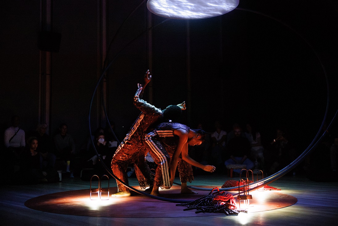 Sweat Variant, Okwui Okpokwasili and Peter Born: let slip, hold sway, 2025. Photography Maria Baranova. Image courtesy the artist and The Whitney Museum of American Art. | Summer at IMMA 2025 | Sunday 1 June  – Sunday 31 August 2025 | IMMA | Image: Sweat Variant, Okwui Okpokwasili and Peter Born: let slip, hold sway, 2025. Photography Maria Baranova. Image courtesy the artist and The Whitney Museum of American Art. | two figures performing in a dark space (though there are hints of spectators in the background); two figures wearing what may be red tracksuits, though grotty ones, are interacting, in that one is leaning backwards, hands in the air, onto the back of the other, who is bent forward as though tending to a fire, though the firre is apparently some arrangement of metal and maybe rope, red; strong lights pick out parts o fthe scene, and there appears to be a circular cloudscape projected onto a surface above the figures 