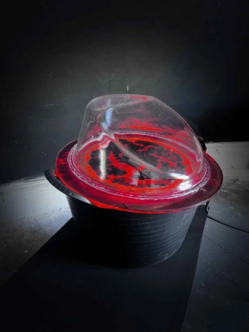 NCAD MFA Fine Art | Saturday 7 June  – Saturday 14 June 2025 | Rua Red | Image: photo, but it is hard to make out what it is of; looks like an upside-down bowler hat with a red plate on it with a clear plastic sort-of hat on taht; the ’plate’ has some sort of black pattern on it, somewhat brainlike; the light soruce is behind the hat 