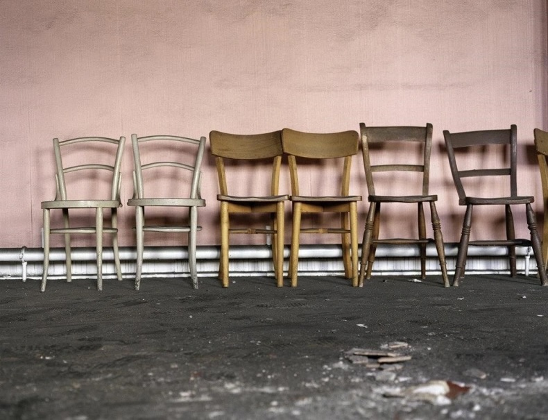 What does it mean to know? | Thursday 12 June  – Saturday 9 August 2025 | The LAB | Image: a photo of seven wooden chairs against a somewhat grubby pale-pink wall; we are viewing them ‘face-one’ at seat level, and the chairs are ranged from left to right across the photo, sitting on a dark-grey floor of some sort, perhaps carpet; we also see white pipes behind the chairs, just above the floor, behind the chairs; the first two chairs are white-ish, the next two yellowish wood, the next two grey-brown wood, the last one like the third and fourth ones, but severely cropped off because at the right edge of the photo; something has been smashed on the floor near the camera 