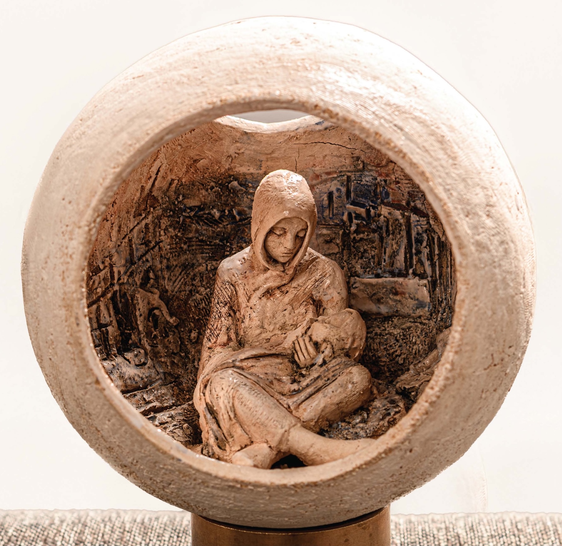 Ann Meldon Hugh: Fragile World, ceramic and bronze, 13 x 13 x 24cm | Ann Meldon Hugh: Fragile World | Wednesday 25 June  – Saturday 26 July 2025 | Toradh2 Gallery | Image: Ann Meldon Hugh: Fragile World, ceramic and bronze, 13 x 13 x 24cm | photo of the artwork; we see a mother breastfeeding a child inside a sphere that is open at the front and above towards the back; the mother is wearing a hooded garment and is looking down at the child; the outside of the globe is rough in texture and off-white; the interior in which the mother is sitting is rougher still, and resembles a blackened cityscape 