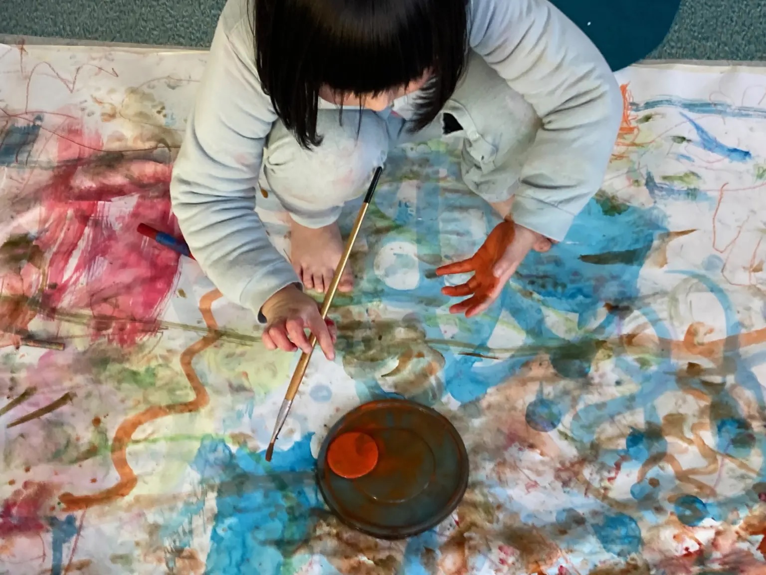 Boghanna Báistí Beaga | Friday 27 June  – Sunday 28 September 2025 | Hugh Lane Gallery | Image: photo from above and in front of a child holding a paintbrush while squatting on a large white cloth that is covered in painted squiggles and other marks; black straight hair, bare feet, white clothes 