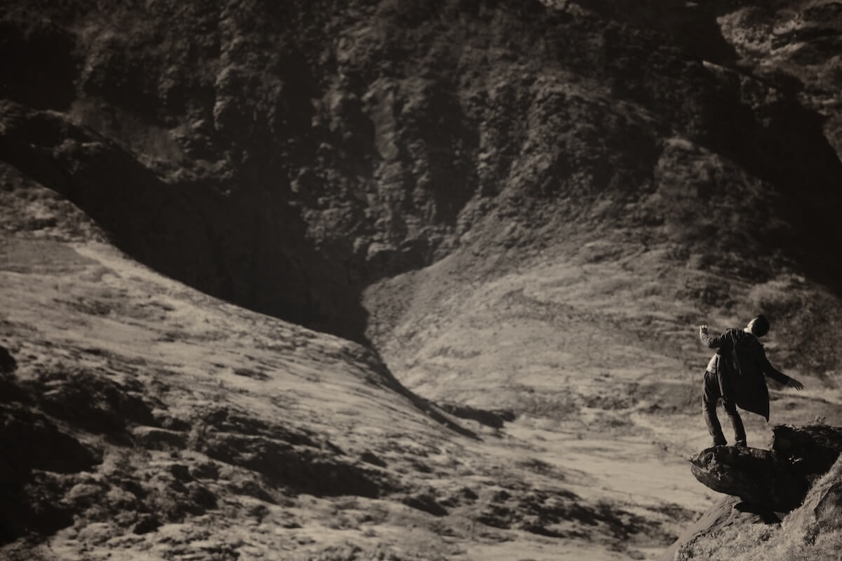 Clare Langan: Elizium, 2014 | Drawing With Light | Saturday 5 July  – Monday 28 July 2025 | SO Fine Art Editions | Image: Clare Langan: Elizium, 2014 | greyscale photo or photo-like image of a figure in a bleak, bare-hills landscape (possible Iceland?); the figure is off to one side on an outcrop above what seems would be a serious drop; the figure is leaning to the right, dark-haired and wearing a cloak – operatic buzz 