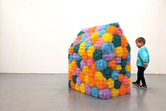 Jane Hayes: Sweet Home Sweet | Tuesday 3 June  – Saturday 23 August 2025 | Garter Lane Arts Centre | Image: photo, of a house-shaped object in what is presumably a gallery space; the house is made of cage-like material and filled, apparently, by large balls of knitting fabric in bright colours; a child, aged 2 to 3, is standing near the house, looking at it 