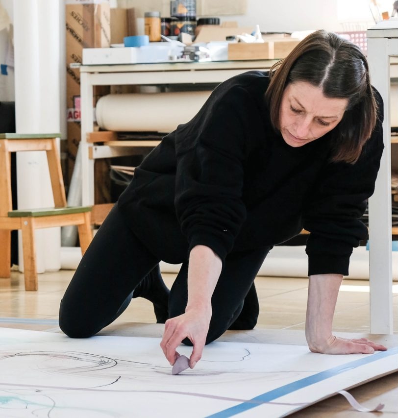 Photography by: Claudio Nego, Photojournalist (for Wexford Arts Centre) | Órla Bates: Sync Shift | Saturday 14 June  – Saturday 26 July 2025 | Wexford Arts Centre | image: a photo, presumably of Órla Bates, on hands and knees in black top and jeans, drawing on what may be paper or canvas that is lying on the floor; the shot is from in front of the artist, who is looking down at the mark being made by what may be a chunk of pigment of some sort, purplish; behind we seem to see an artist’s studio, and possibly very large rolls of paper 