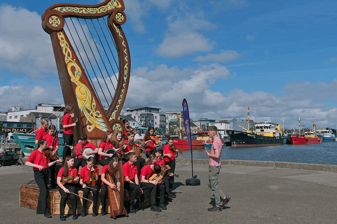 In the Moment: Capturing the Fleadh 2024 | Saturday 2 August  – Wednesday 13 August 2025 | Wexford Arts Centre | Image: the photo, taken on a waterfront pier, shows a group of young (probably teen) musicians in red tops and dark trousers holding various instruments as they attend to their director, apparently equally young, standing in front of them in a pink t-shirt and grey trousers; towering over them is a Celtic harp with Celtic design; in the background we see buildings and ships; it's sunny with a bit of cloud 