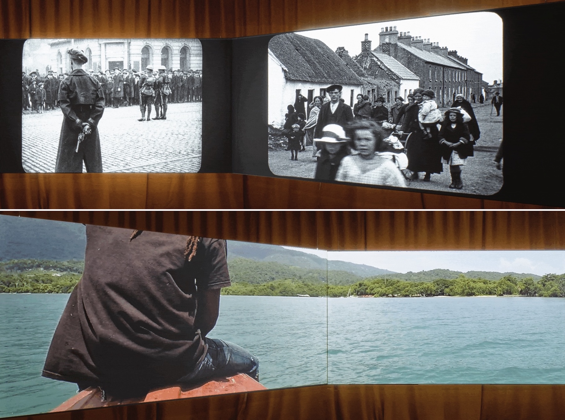 Marianne Keating: An Ciúnas / The Silence | Tuesday 26 August  – Wednesday 1 October 2025 | Wexford Arts Centre | Image: still image from two projections in an art space or similar; each projection is on two surfaces at an angle to each other; top projection contains two images, both black-and-white; on the left there is a soldier near us, back turned to us, holding revolver behind back; cobbled street, suggestion of tram line; two more soldiers closer to crowd facing the soldiers; men, women, some kids; fairly fancy building behind; in second photo of top image, a street scene with collection of people of many ages including very young – possibly extended family; thatched cottage on left, that they are passing, derelict house next to it, then another bungalow, then terrace of houses; neighbour appears to be watching from thatched cottage door; bottom image is possibly a video projection; we’re in the Caribbean? – sea, trees in the distance; photographer is probably in small boat, with someone sitting on prow; we just see that persons’s back and a leg and arm, plus hint of dreadlocks