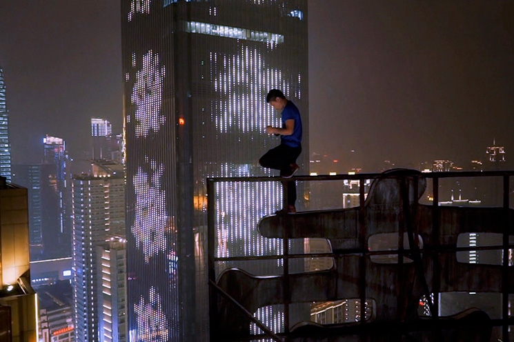 Still from 'Nightworld', 2019, directed by Adam Smith | City as Character Film Programme | Saturday 9 August  – Sunday 26 October 2025 | Butler Gallery | Image: Still from 'Nightworld', 2019, directed by Adam Smith | someone, apparently, standing precariously on a railing at the edge of (presumably) a skyscraper; they appear to be on their phone; behind them we see many skyscrapers in a misty-night city, probably Chongqing 