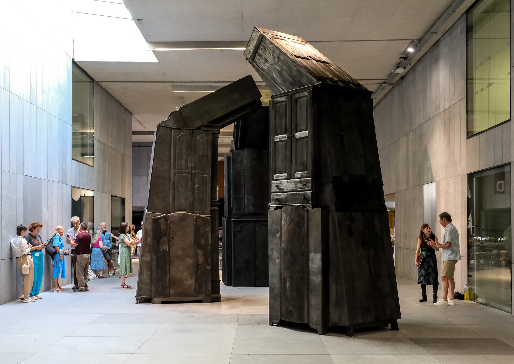 Nina McGowan: Container | Thursday 14 August  – Friday 19 September 2025 |  | Image: installation photo; we see a huge sculpture in a large atrium; it seems to be in three (or more) parts, and seems to consist of many wardrobes / closets merged together to form towering shapes with the top wardrobe / closet leaning inwards towards the other similar assemblages; they are the colour of streaked / mottled / blemished creosote; we see people around the edges of this work; the space is modern, probably the council offices 