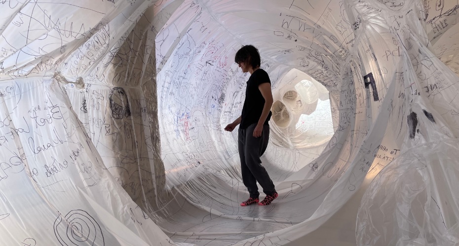 Olga Diego: Metamorphosis | Monday 6 October  – Friday 10 October 2025 |  | Image: photo of a dark-clad figure standing inside a sort of tubular, loose-fitting polytunnel; through the semi-transparent skin of the tunnel we can see that the external surface of the tunnel is covered in writing and possibly also some drawings; there are strange shapes further down the tunnel, behind the figure, and we see the shadow of a column exterior to the tunnel 