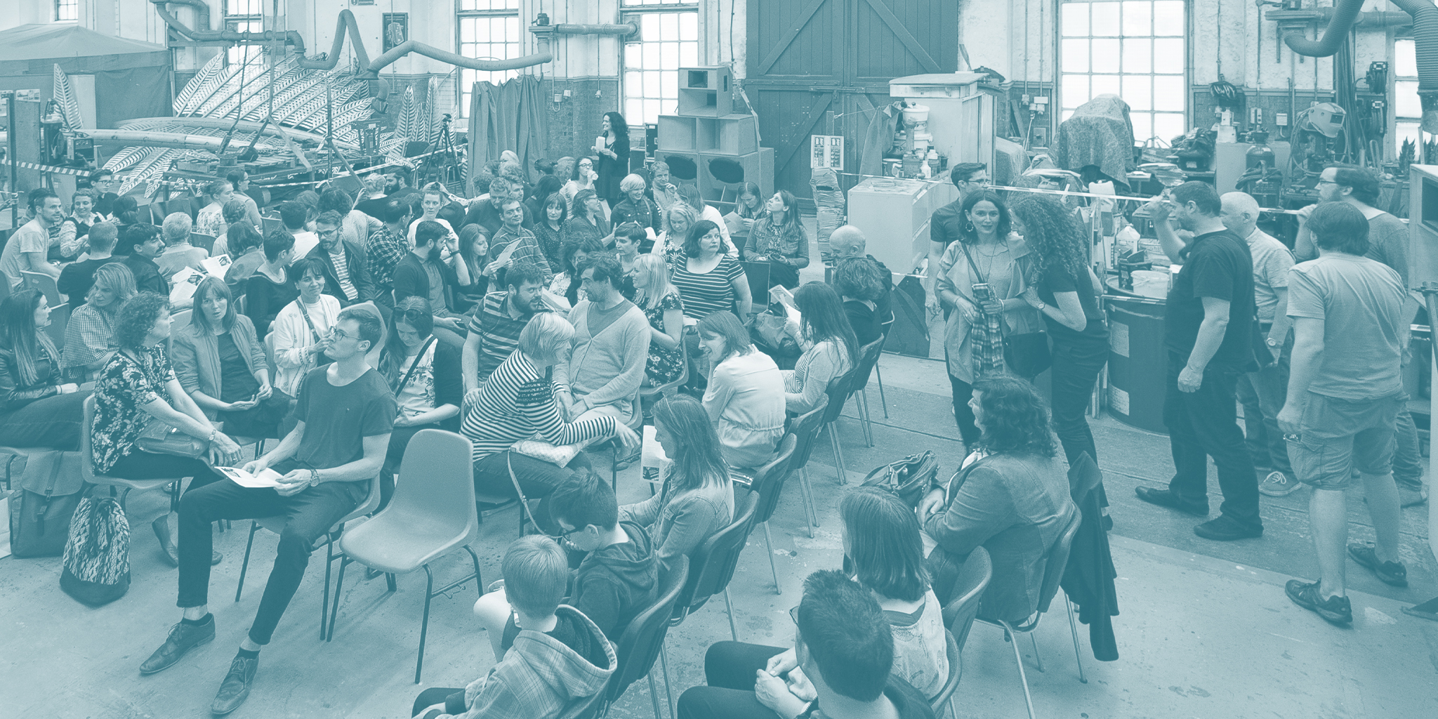 Career of an Artist | Tuesday 4 November  – Tuesday 2 December 2025 | National Sculpture Factory | Image: photo view of NSF floor, in slightly blue-ish greyscale; we see a lot of people sitting on chairs having the chats; ther are bits of works in progress to be seen in the background 