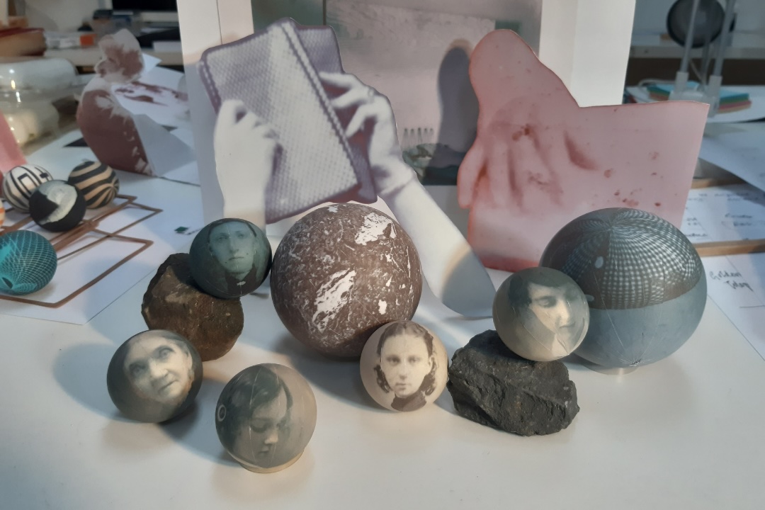 courtesy of Susan MacWilliam | Susan MacWilliam: Table Turning | Thursday 20 November 2025  – Saturday 21 February 2026 | Ormston House | Image: courtesy of Susan MacWilliam | photo; we see seven spheres on a tabletop; five of them are smaller than the remaining two, and bear photos on their surfaces – black-and-white, old-looking; one of the other spheres is made from stone, the other is reflective; two ’ordinary’ stones are also in the mix; oh, and there are four further smaller spheres off to the left, but they bear no photos; behind the nearer spheres are what appear to be paper or card cutouts of hands touching or holding things; there seems to be another like this farther away; this is probably a studio space  