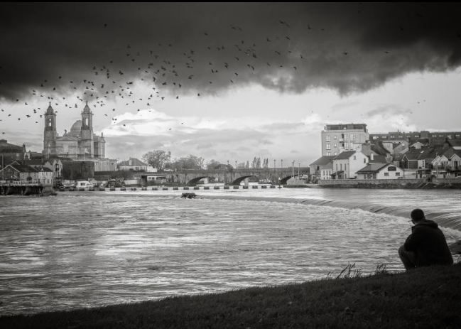Moments in Focus: Athlone Photography Club Exhibition | Saturday 29 November 2025  – Monday 2 February 2026 | Luan Gallery | Photo Credit: Under A Cloud by Stephanie Reddin | greyscale photo of view of the Shannon in Athlone from Burgess Park looking northwest across the weir and towards the old bridge; someone i sitting at the edge of the right towards bottom right of the photo; we see a swarm of birds under a dark, cloudy sky, though it’s a brighter day in the distance. 