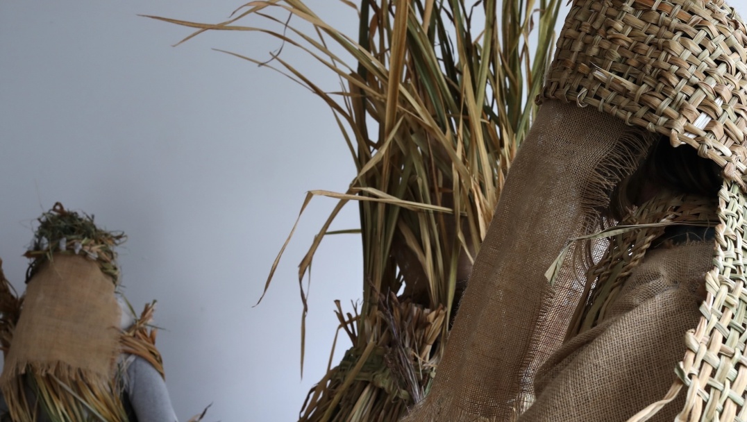 ^: Waking the Land | Saturday 22 November 2025  – Saturday 14 February 2026 | The Dock | Image: photo – we see a sheaf of grass of some sort quite close to the camera, and it seems that it may have provided the material for two woven strips that we see towards top right and right in the photo; there seems to be more sheafery hidden behind burlap or equivalent, and farther away we may be seeing someone wearing this gear, along with some grass and stones, maybe, on their head; a grey wall behind 