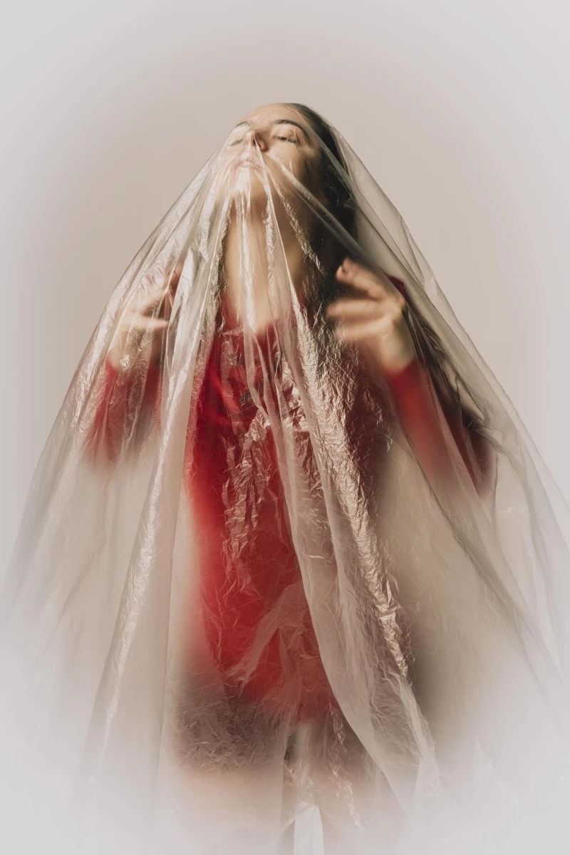 Patient Labyrinth. Photo by Fionn McCann | Junk Ensemble: Patient Labyrinth | Friday 12 December 2025  – Saturday 7 February 2026 | Rua Red | Image: Patient Labyrinth. Photo by Fionn McCann | we see a figure’s upper body, long black hair, red top, pale skin, hands raised to shoulder height, head raised and looking a bit to our left; the figure is covered by a transparent plastic sheet; white-to-cream background 