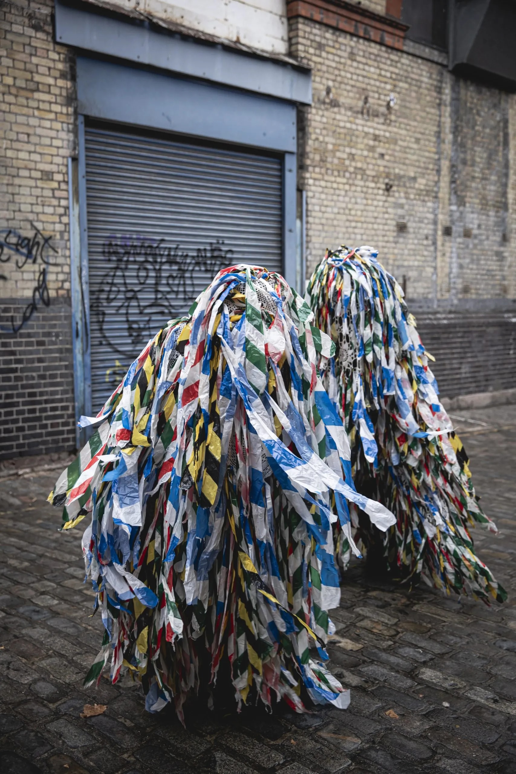 Joe Lang | Lunatraktors: Our Deepest Apologies | Saturday 10 January  – Saturday 24 January 2026 | GOMA Gallery of Modern Art | Image credit: Joe Lang | photo of (presumably) two figures outside on a slightly wet cobbled street; each figure is entirely draped with strips of traffic / warning / safety tape of different colours 