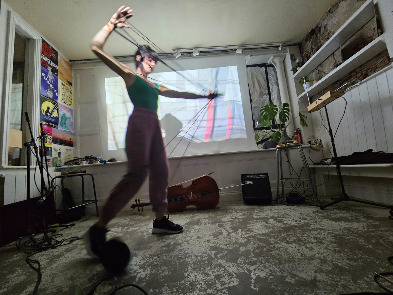 Vicki Davis: Out of the Darkness, There Comes Light | Tuesday 2 December  – Monday 22 December 2025 | Uillinn: West Cork Arts Centre | Image: photo in which we see a figure, presumably the artist, in what seems to be a cross between a music-performance space and a studio; musical equipment, especially possibly a cello lying on its side towards the back wall; the figure seems to be weaving / unwinding / dunno wool or threads, arms wide, in front of a projection onto a sheet or similar on the back wall; the photo is shot from a low angle, almost on the floor; the figure is towards the middle of the image, facing to our right, eyes shut; green sleeveless top and bare arms, purple tracksuit bottom (?), black trainers; and more 