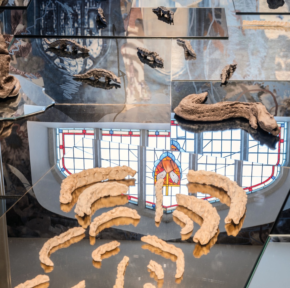 Alice Rekab: Clann Miotlantach / Mythlantics | Friday 30 January  – Sunday 5 April 2026 | Limerick City Gallery | Image: photo, possibly an installation shot; we seem to be looking at many rectangular mirrored surfaces on top of or abutting each other; they seem to be reflecting part of a partially stained-glass window, but also possible artworks; crawling across the mirrors are critters made of clay (probably), reptilian, possibly miniaturised dinosaurs; closer to the camera seem to be partial critters in pale yellow-ochre clay 