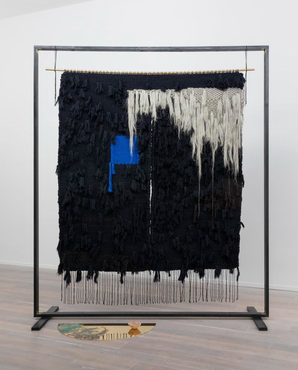 New Work New Artists  | Friday 19 December 2025  – Friday 13 February 2026 | Green On Red Gallery | Image: Emily Waszak  We Speak Through Worlds  2023  Wool, wood, etched brass, clay  280 x 220 x 60cm | photo of a probably large steel frame from which is hanging a slort of blanket – mostly black, a patche of blue in a different texture from the black background, which seems to e relatively smooth, but with protruding ribbon-shapes; ther eis also a fairly large triangular area towards to right of the tapestry that has a cross-hatched pattern of white wool (?), with large trailing areas of the same material; just in front of the frame, a bit to the left when looking face-on, is what seems to be a semi-circular copper sheet, very reflective, with some engraving, possibly, or greenish material on it, and small bowl  