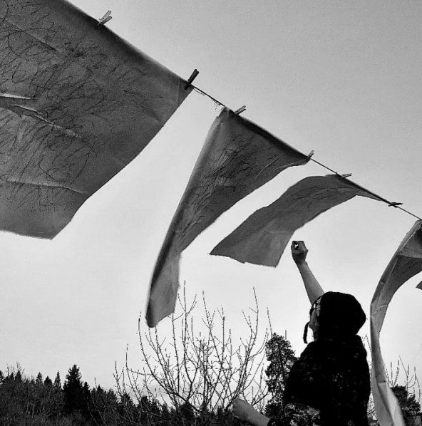 Eileen Fair and Gary Robinson: Nósanna | from Saturday 7 March  | Roscommon Arts Centre | Image: photo, black-and-white; it appears to be of an artist drawing onto pieces of paper or fabric that is clothes-pegged to a clothes-line; they are blowing in the wind, and the artist is stretching up their right arm to draw; we only see from waist up of the artist; conifers and a bare tree or two in the background 