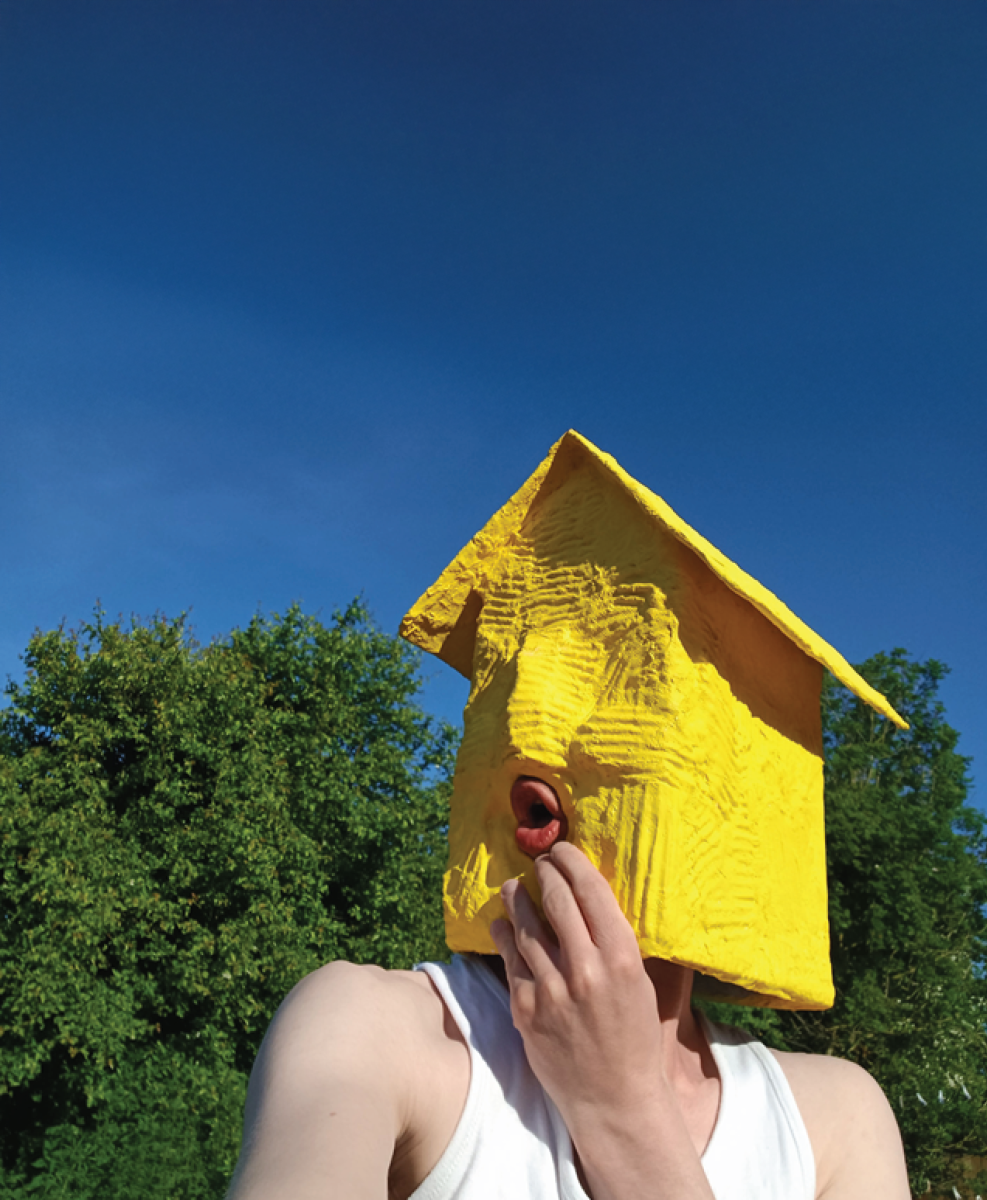 twixt – group exhibition | Wednesday 14 January  – Wednesday 25 March 2026 | Atypical Gallery | Image: photo a figure of whom we just see head and  bare shoulders with white top; the head, though, is a bright yellow funky house with a shape for a nose and a hole for the mouth, in which we see lips making an O; there are trees / bushes behind and a blue sky 