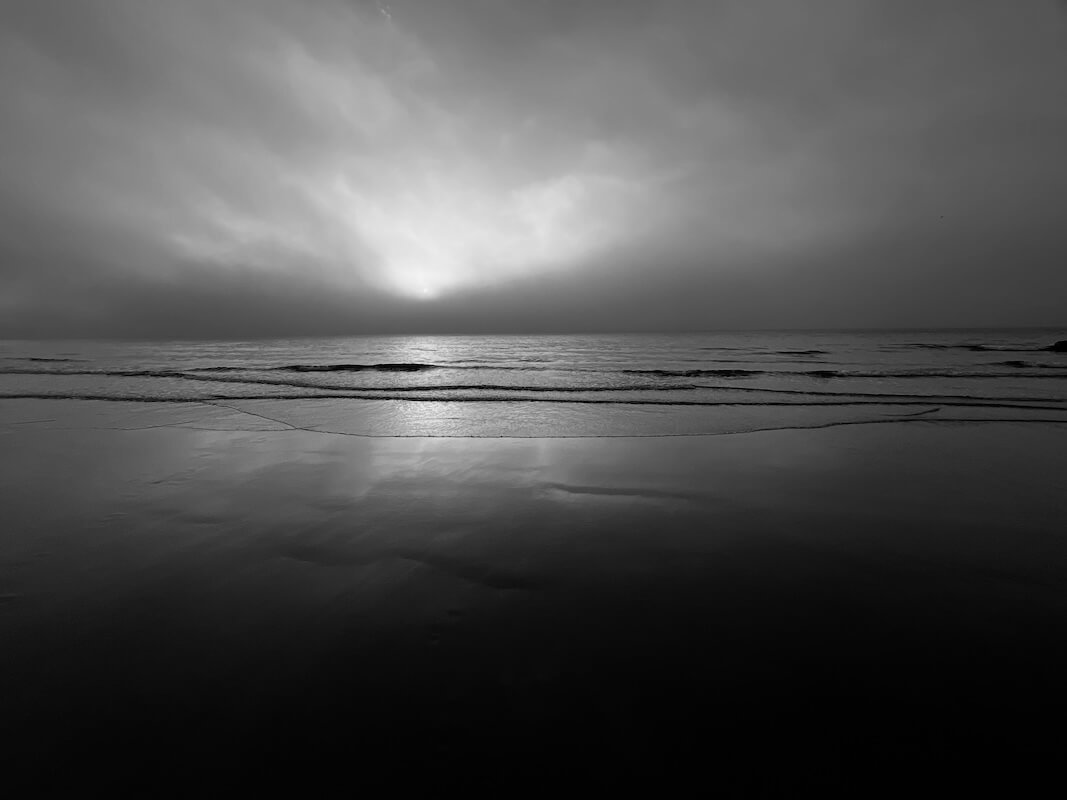 Michael Corrigan: Blainroe, Wicklow I, Archival Pigment Print, ed of 5, 40h x 50w cm | Michael Corrigan: Margins | Thursday 5 March  – Saturday 4 April 2026 | SO Fine Art Editions | Michael Corrigan: Blainroe, Wicklow I, Archival Pigment Print, ed of 5, 40h x 50w cm | black-and-white photo taken standing on a sandy beach, presumably looking east as it is taken in Wicklow and we see a low sun, mostly occluded by clouds, a dark horizon, a calm sea with calm waves rolling towards wet sand reflecting a wispy sky 