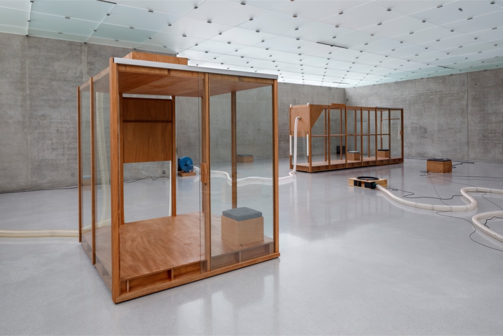 Tarek Atoui: Wind House 1 & 2, installation, Kunsthaus Bregenz (Austria); © Markus Tretter | Tarek Atoui: Souffle Continu, Sunflowers | Saturday 21 February  – Sunday 19 July 2026 | IMMA | Image: Tarek Atoui: Wind House 1 & 2, installation, Kunsthaus Bregenz (Austria); © Markus Tretter | photo of two ‘houses’ in an exhibition space; each is made of wood, not very big, some glass or perspex, gaps; geometrical; the farther house, which is mostly framework, seems to have some sort of white suction hose attached to it