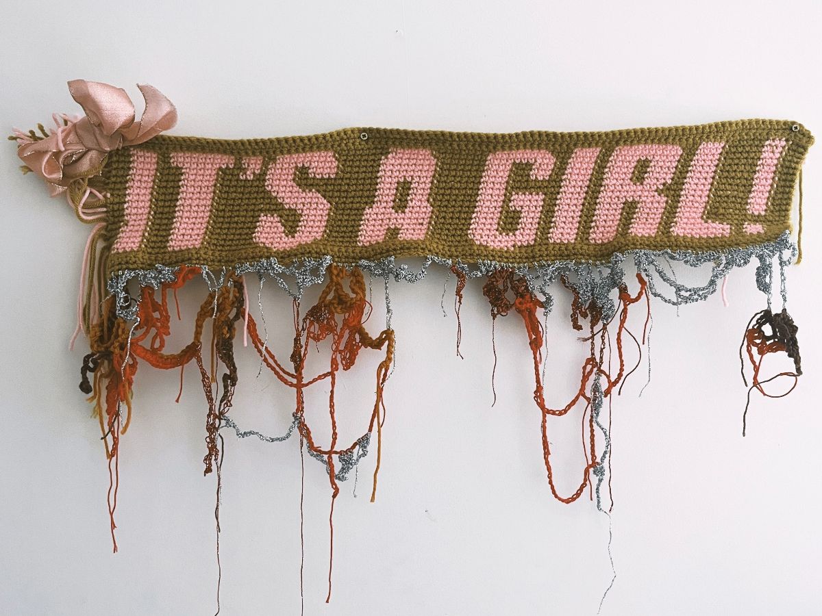 Shirani Bolle, It’s a Girl, 2025. Yarn, thread, ribbon; dimensions variable. Courtesy of the artist | Shirani Bolle: this has nothing to do with me | from Saturday 14 February  | SIRIUS | Shirani Bolle, It’s a Girl, 2025. Yarn, thread, ribbon; dimensions variable. Courtesy of the artist | photo of a scarf, or scarf-like tapestry object, screwed to a wall; it’s background is an olive green, on which, or into which, the words ‘IT’S A GIRL’ is woven / crocheted in a pale pink; there is a sort of one-complex-flower bouquet top left on the scarf, and random skeins of thread and wool hang and loop down from the bottom edge of the scarf; some of these appear to be silver 