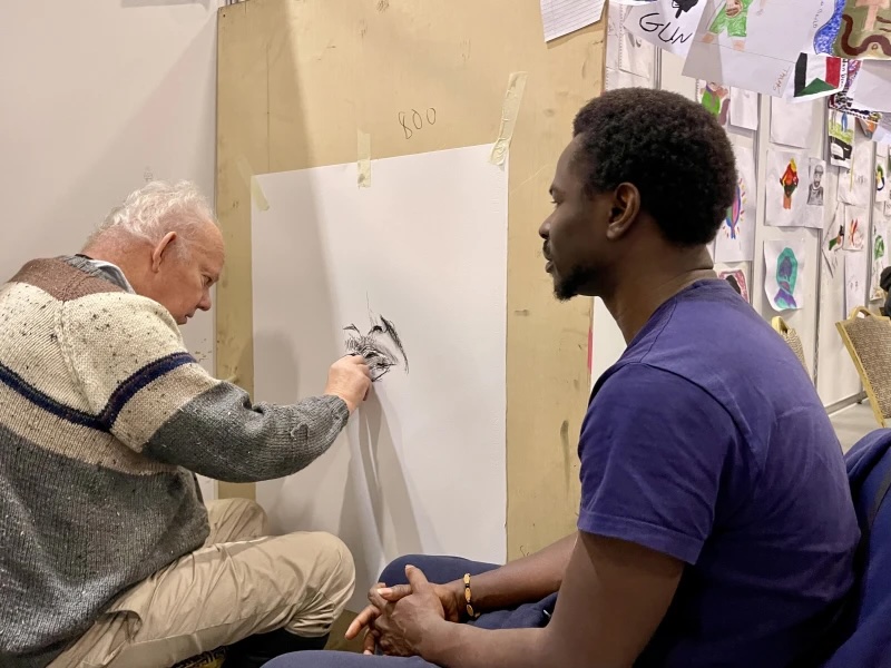 Brian Maguire: These Beautiful Men | Friday 6 March  – Saturday 9 May 2026 | Rua Red | Photo of the artist drawing a portrait – the artist to the left, seen sideways from behind, and the sitter sitting on the right, sitting in profile 