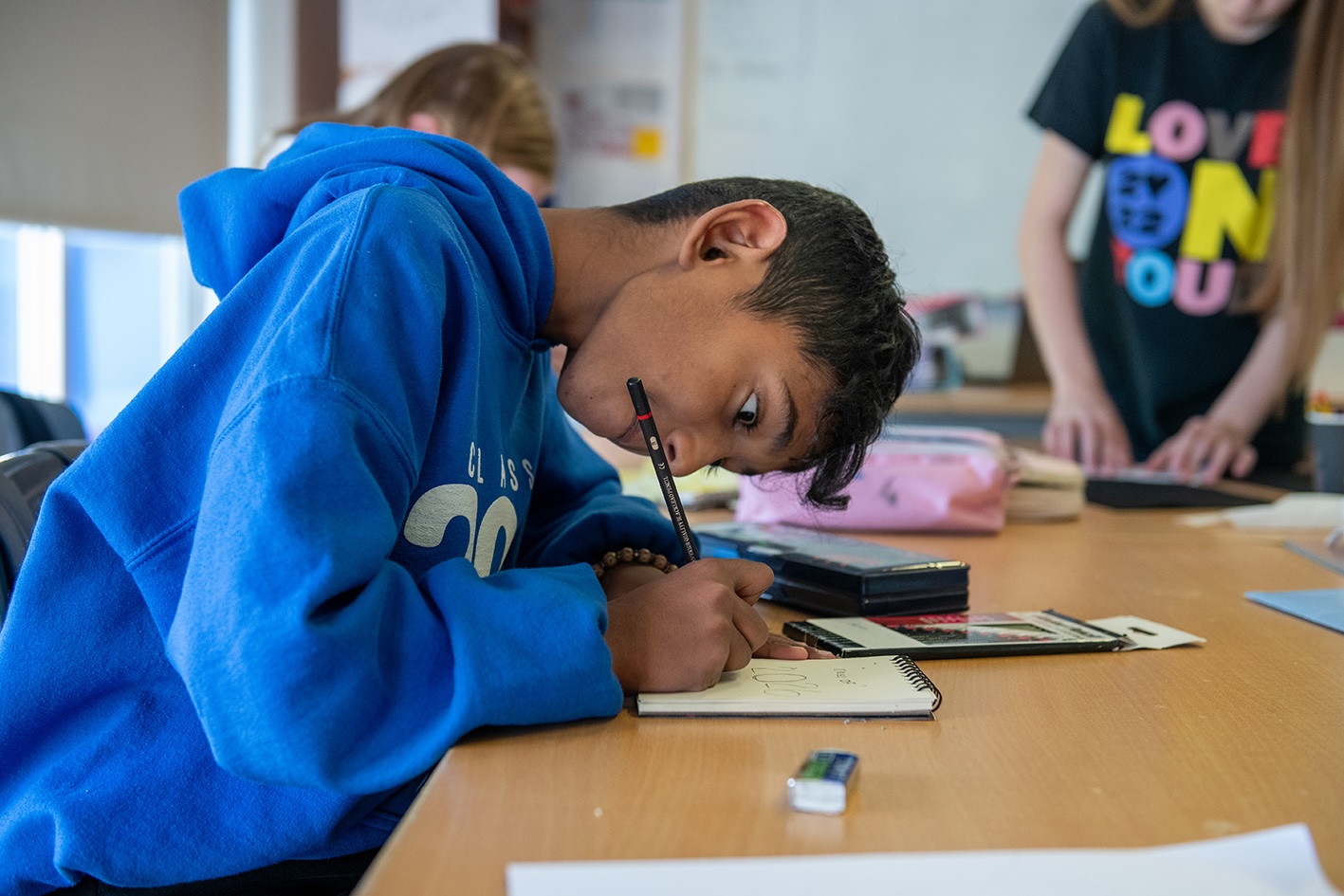 Living Arts Project 2026 | Tuesday 31 March  – Thursday 30 April 2026 | Wexford Arts Centre | photo: A pupil of Castlebridge NS | side-on shot of a kid who looks really into drawing on a small-ish drawing pad; bent over it, intent, intense; bright blue hoodie; side-on photo; glimpses of two others in the background 