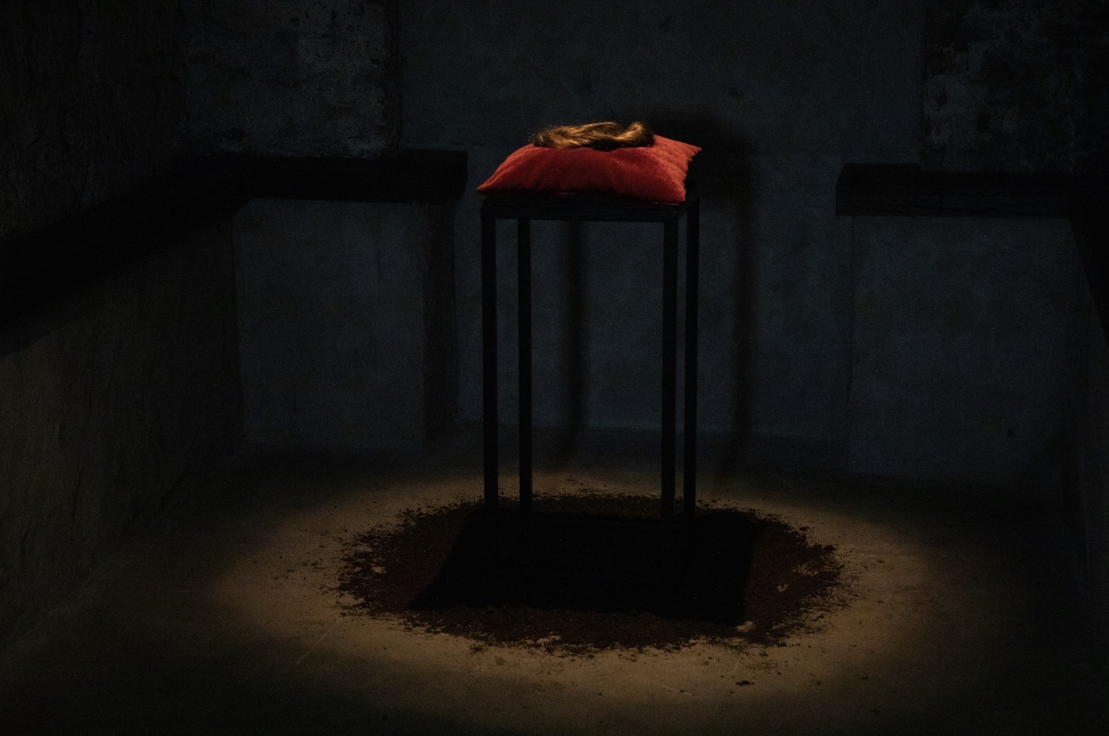 Lara Quinn | Sanctum | Group Exhibition | Thursday 2 April  – Saturday 25 April 2026 | GOMA Gallery of Modern Art | Image: Lara Quinn | this appears to be a photo of brunette wig on a red cushion on a very tall black table with a top just wide enough to hold the cushion, all on a black circle on the ground, possibly made of dirt, with a halo of light around it on the floor, then some aspects of the darkened room; a light is shining down from above, centred on the wig 