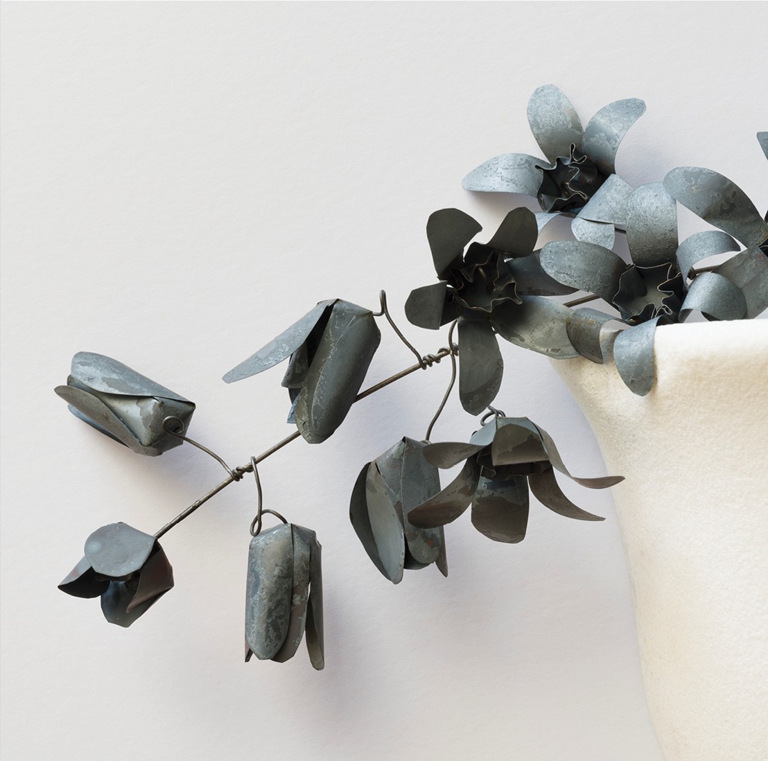 detail: Mary Nagle, Oblatio, marble, fired steel, 15 x 31 x 6cm. Photo credit: Roland Paschhoff | Mary Nagle: MURMURARE | Friday 27 March  – Sunday 3 May 2026 | Limerick City Gallery | Detail: Mary Nagle, Oblatio, marble, fired steel, 15 x 31 x 6cm. Photo credit: Roland Paschhoff | photo of a stem of flowers, tulip-like, sticking sideways and downwards across the image, from a white container of some sort, of which we see just its left side; the flowers are in metal, metallic grey, and are bound to the stem by grey wire; the wall behind is white 