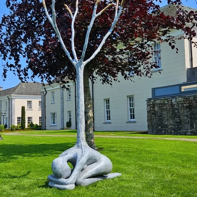 Orla de Brí, Source, bronze, unique, 270 x 127 x 87cm | Site Unseen: Garden & Outdoor Sculpture | Thursday 30 April  – Saturday 23 May 2026 | Solomon Fine Art | Orla de Brí, Source, bronze, unique, 270 x 127 x 87cm | the photo captures a sculpture of a curled-up figure with a leafless tree growing out of their back; the bronze is patinated to seem closer to silver, though at the tops of the branches it looks more like gold; the sculpture is on a tidily mown lawn, and behind it (in terms of where the photographer is standing) there is a tree full of dark-red leaves, and the effect is that has leaves; it is sunny, and a grand house looks out over the lawn 