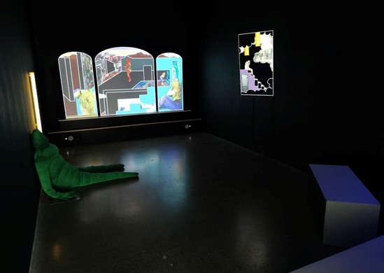 Sorcha Browning | BA Visual Art Graduate Exhibition | Saturday 23 May  – Saturday 13 June 2026 | Uillinn: West Cork Arts Centre | Image: Sorcha Browning, Eden, Image courtesy the artist | photo of an installation in a small-ish (probably) room with black walls; a stuffed all-green, mostly featureless figure is sitting on the floor and leaning against the wall on the left; a bit farther along the left wall is a vertical light tube; on the wall at the end there is a sort of triptych in the shape of a make-up mirror or medieval altar decoration in a church; in its panels we see a blonde, long-haired figure in at a few points in a geometric-ish space in funky colours; it’s a sort of photo collage, backlit; on the right wall is apparently a brigly electronic painting of some sort – geometric shapes in black, yellow, white, a table and chair in clouds… 