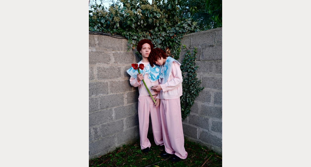 Merve Sagit, Between Sisters, 2026, Photographic print, 60 × 84 cm, Image courtesy of the artist. | Resolve 26 | Tuesday 26 May  – Sunday 7 June 2026 | Royal Hibernian Academy | Image: Merve Sagit, Between Sisters, 2026, Photographic print, 60 × 84 cm, Image courtesy of the artist. | we see two figures, presumably the eponymous sisters, standing in the corner of what is probably a garden; one system is holding the stem of two large red flowers; both are dressed the same – pink outfits with blue toppings; the garden walls are high and of raw concrete blocks, with ivy clambering over; one sister is leaning into the other 