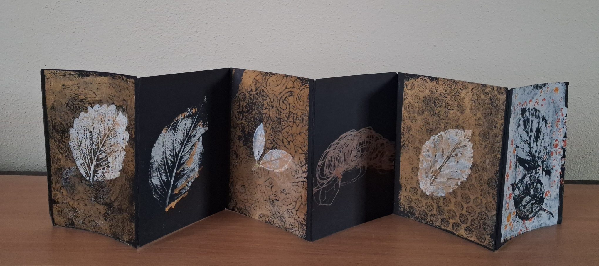 Bealtaine 2026 Exhibition | Friday 1 May  – Sunday 31 May 2026 | Uillinn: West Cork Arts Centre | photo of a folding card – six images face us as the card stands on a wooden surface; the base colour of each image is black, with a pattern in brown or gold sometimes over it; then there is a central item depicted, usually a leaf or two, though one is just a swirling 