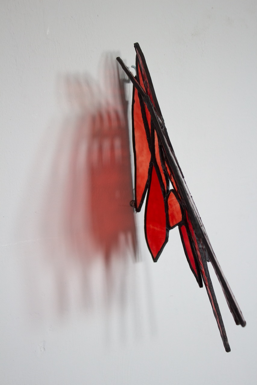 Oisín Tozer: Danus (Red), 2026, foiled stained glass, 30 x 20 x 8cm, photo : Jan McCullough | Oisín Tozer: Breathing in the Dark | Friday 22 May  – Friday 3 July 2026 | Green On Red Gallery | Oisín Tozer: Danus (Red), 2026, foiled stained glass, 30 x 20 x 8cm, photo : Jan McCullough | the artwork is viewed from the side; viewed this way, it looks more like an insect, possibly a buterfly, resting on the wall; the light sources create a multiplicity of shadows 