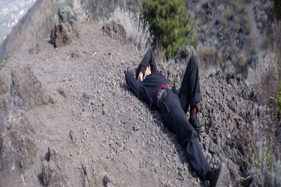 Rebecca Moccia | Rebecca Moccia: Nostalgism | Friday 15 May  – Sunday 28 June 2026 | Temple Bar Gallery + Studios | Rebecca Moccia | tilt-shift, possibly, photo or still of a figure lying on the ground in a pebbly, rocky, dry landscape; the figure is shielding their face, possibly from the sun, with one arm, while the other appears to be supporting the head; the left leg is raised and bent at the knee, the right leg stretched fully, from roughly the centre of the photo to close to the bottom right of the image; we also see some vegetation, and there is a hint of a road in the distance 