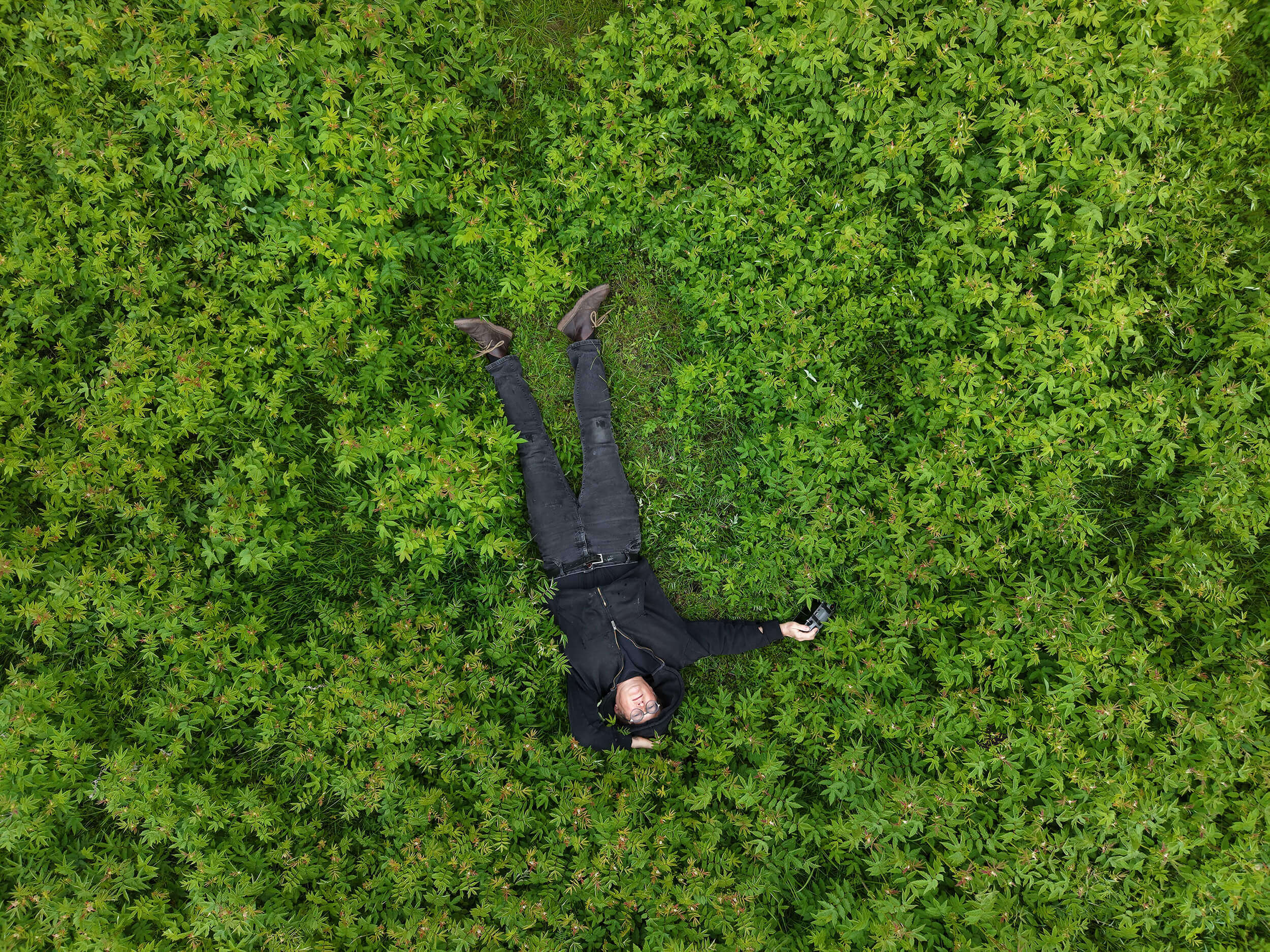 Amanda Ralph: Field Station: Cornafulla | Wednesday 1 July  – Sunday 30 August 2026 | Luan Gallery | Aerial photo of a figure – probably the artist – lying in a field (probably) of grass and other green growth; the figure is upside-down, in the sense that the head is towards the bottom of the image and the feet towards the top; dark top and jeans, brown shoes 