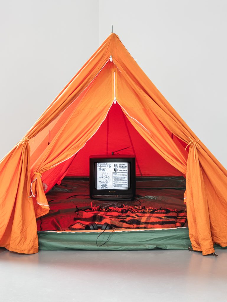 Brian Duggan | Memory of a Free Festival | Saturday 8 August  – Thursday 3 September 2026 | Wexford Arts Centre | Brian Duggan | photo; we are looking directly at the entrance to an orange tent within, apparently, a slightly larger orange tent; both tents have their zipped-open front sides tied open; on the floor of the inner tent we see a television with aerial; the screen is lit up, and on it we seem to be seeing the original leaflet for ‘Ireland’s first anti-nuclear show’, with logo, map, and details; there is a blanket on the tent floor just inside the entrance, and headphones 