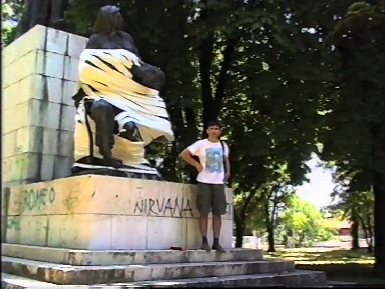 Siniša Labrović, Bandaging a Wounded Man, 2000, Video documentation of action, Video, sound 22:55 min, loop, Camera: Emil Tomasović, Artist’s assistant: Damir Šodan,© Siniša Labrović, Museum of Fine Arts, Split, Croatia. | Technologies of Peace: Art and Transition in Contemporary Europe (1989-2026) | Saturday 12 September 2026  – Sunday 9 May 2027 | IMMA | Siniša Labrović, Bandaging a Wounded Man, 2000, Video documentation of action, Video, sound 22:55 min, loop, Camera: Emil Tomasović, Artist’s assistant: Damir Šodan,© Siniša Labrović, Museum of Fine Arts, Split, Croatia. | location is possibly an urban park; we see a statue on a chunky, somewhat raised plinth; the statue may depict a mother holding across her lap the body of a fallen soldier; however, much of the statue is covered by white tape or toilet roll; the plinth bears the word ‘ROMEO” in green, and ‘NIRVANA’ in black; a figure is posing in fron of the statue, in t-shirt and shorts 